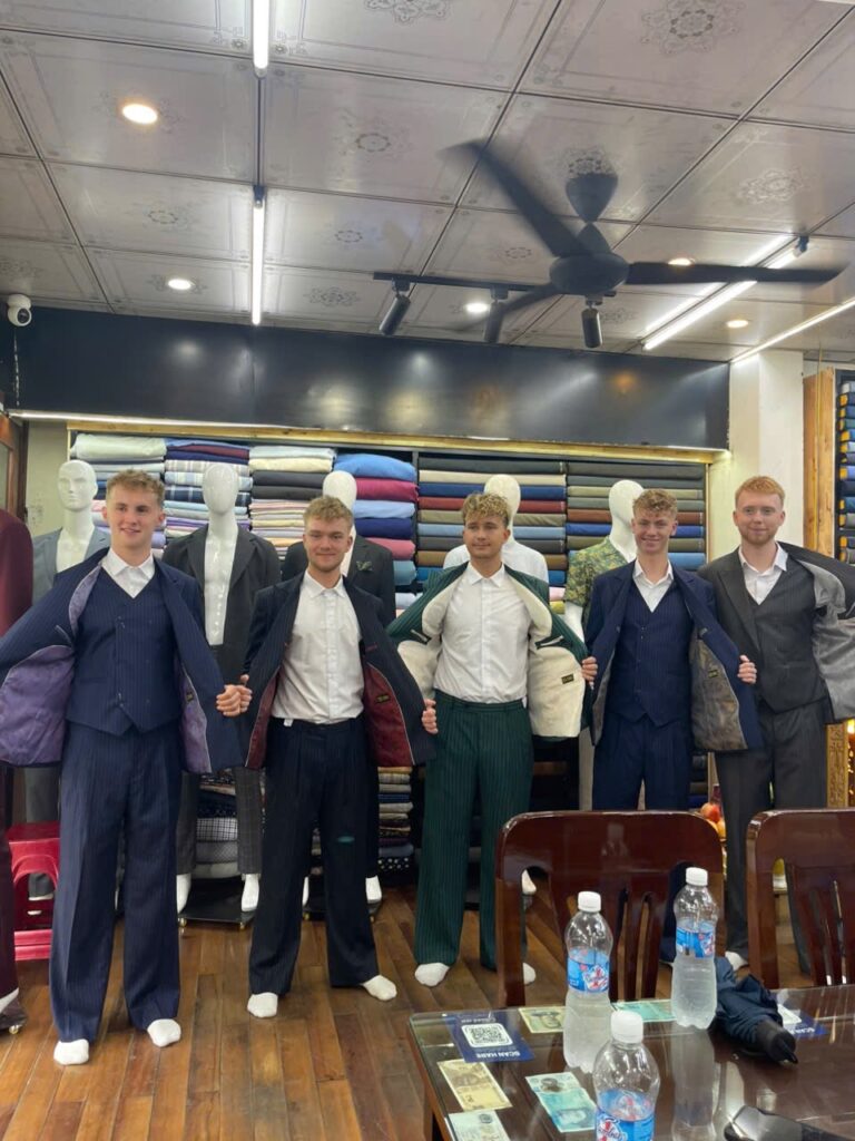 Group of customers wearing custom tailored suits at Trung Duc Tailor shop in Hoi An Vietnam showing handmade suits and premium fabrics.